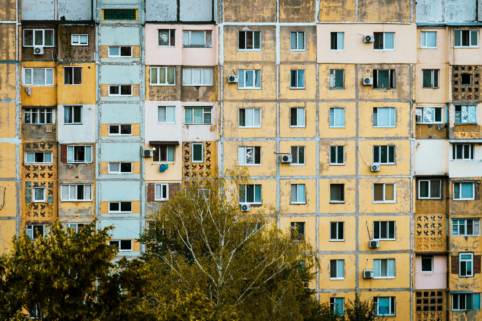 Жёлтая панелька в утреннем свете Фасад многоэтажного панельного дома в тёплых жёлтых и бежевых тонах, окна с кондиционерами и решётчатыми балконами, на переднем плане кроны деревьев.